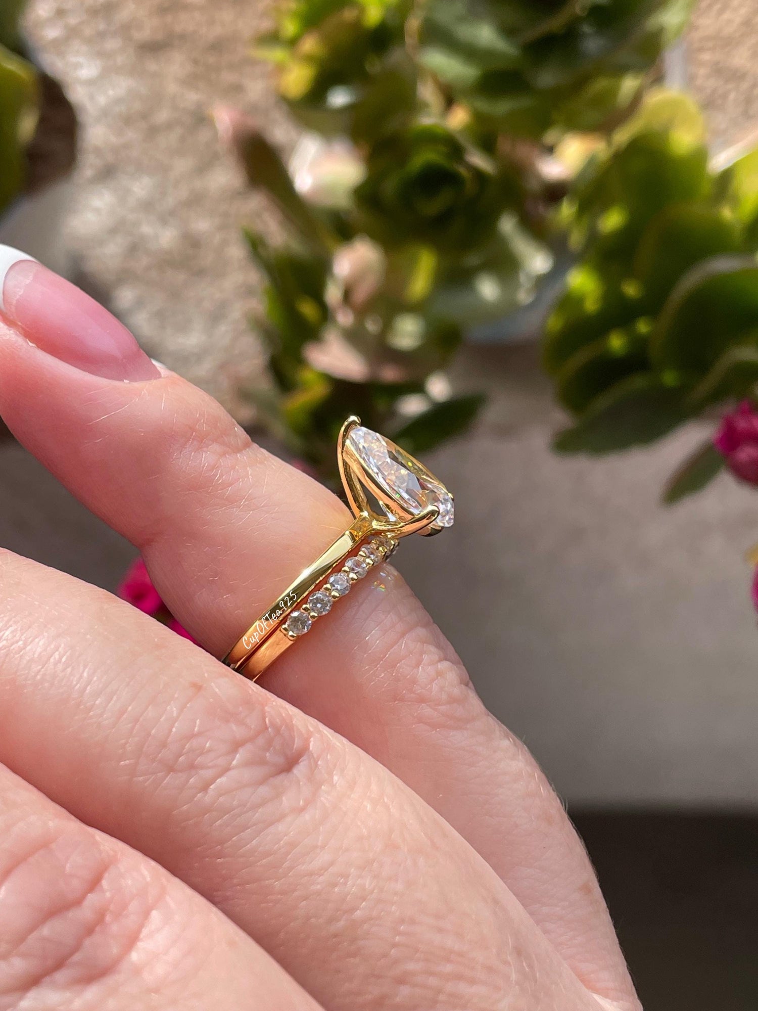 May include: A gold pear-shaped engagement ring with a pave band. The ring is set with a large, clear pear-shaped diamond. The band is set with smaller diamonds. The ring is shown on a finger.