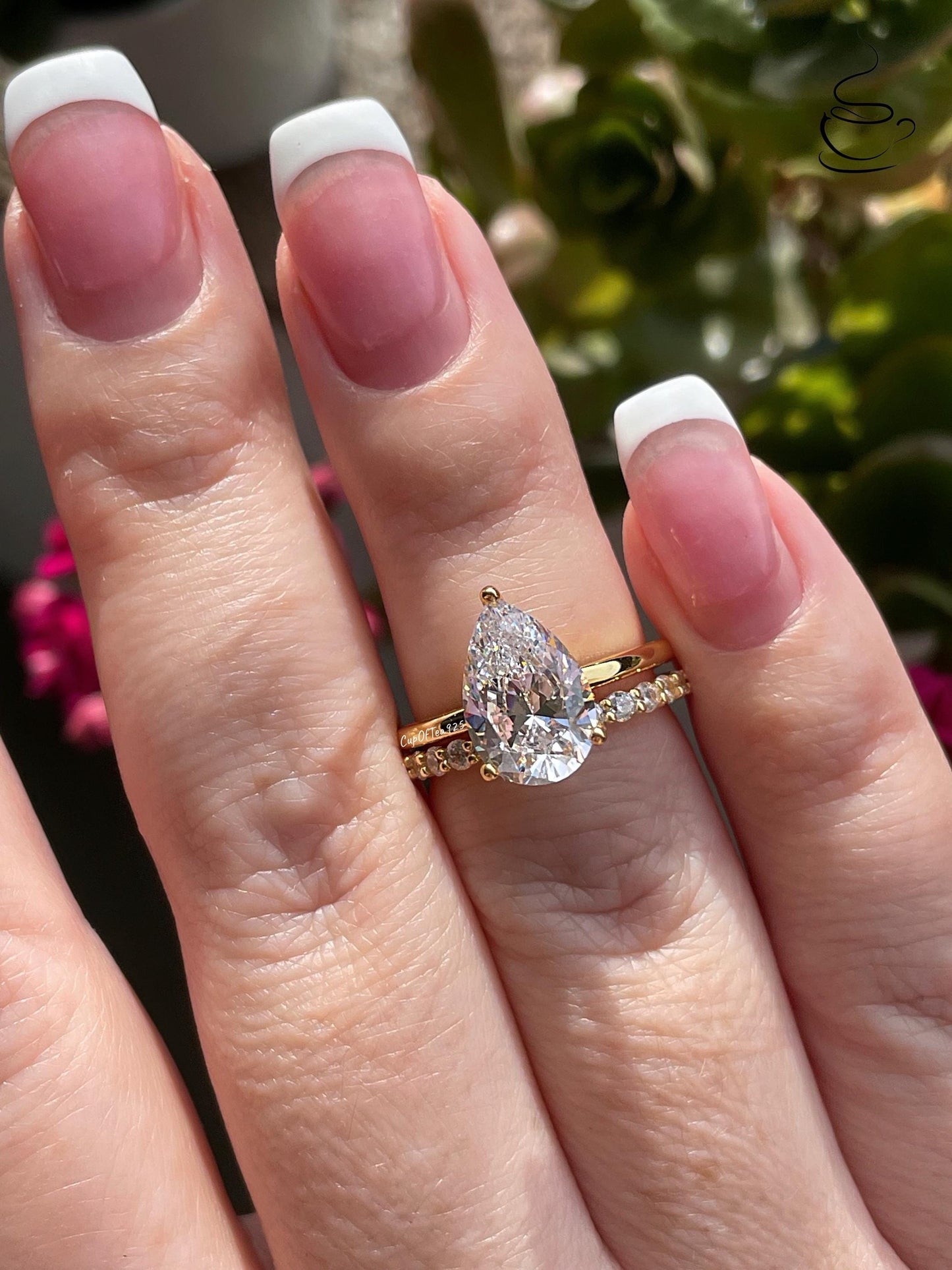 May include: A pear-shaped diamond engagement ring with a gold band and a smaller diamond band. The ring is set with a large, clear pear-shaped diamond in the center. The smaller band is set with smaller diamonds that are arranged in a pave setting.