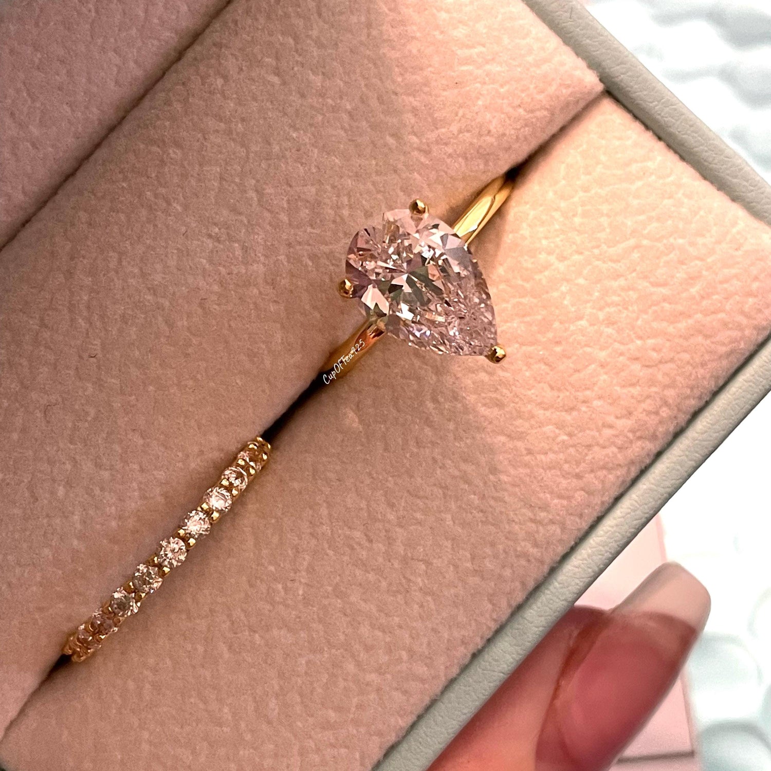May include: A pear-shaped diamond engagement ring with a gold band. The ring is in a light blue ring box with a white interior. The diamond is clear and sparkly.